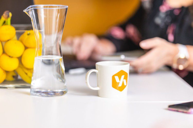 Kahvikuppi, jossa on Verkkoaseman logo, pöydällä vesikarahvin vieressä